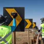 Duas pessoas com coletes refletivos junto a placas de sinalização em uma rodovia