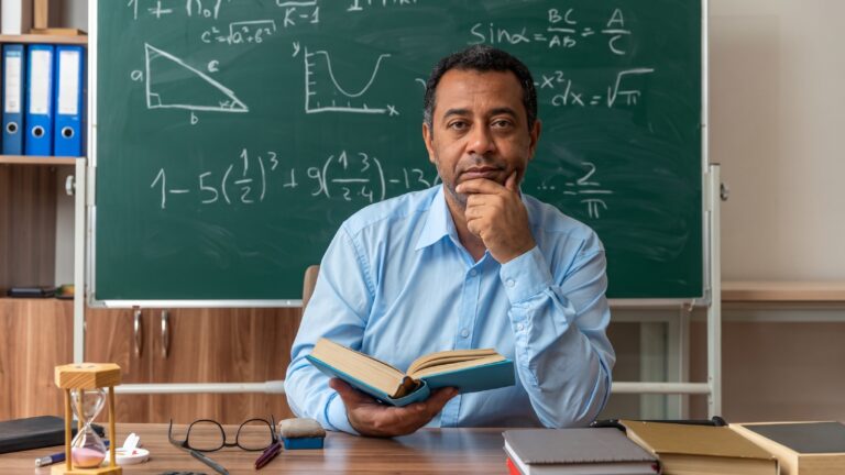 Professor sentado à mesa em sala de aula, segurando um livro aberto, diante de um quadro-negro com fórmulas de matemática
